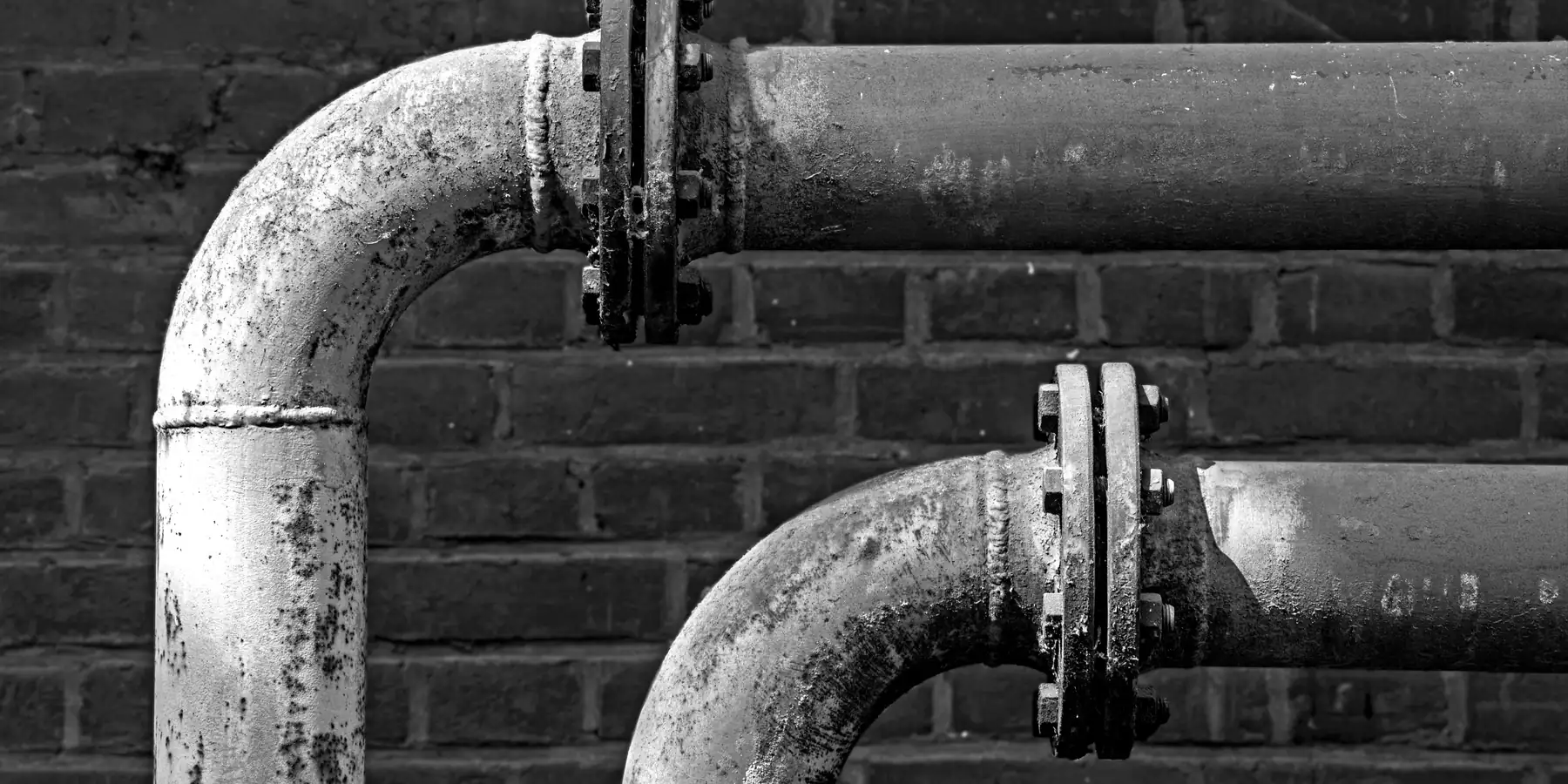 Two rusted pipes with flanges bending sharply against a dark brick wall