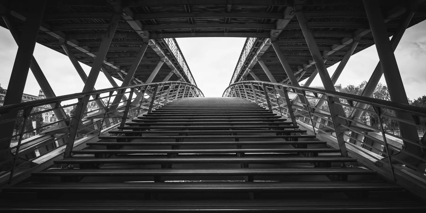 Steel footbridge staircase rising between converging girders toward a bright opening