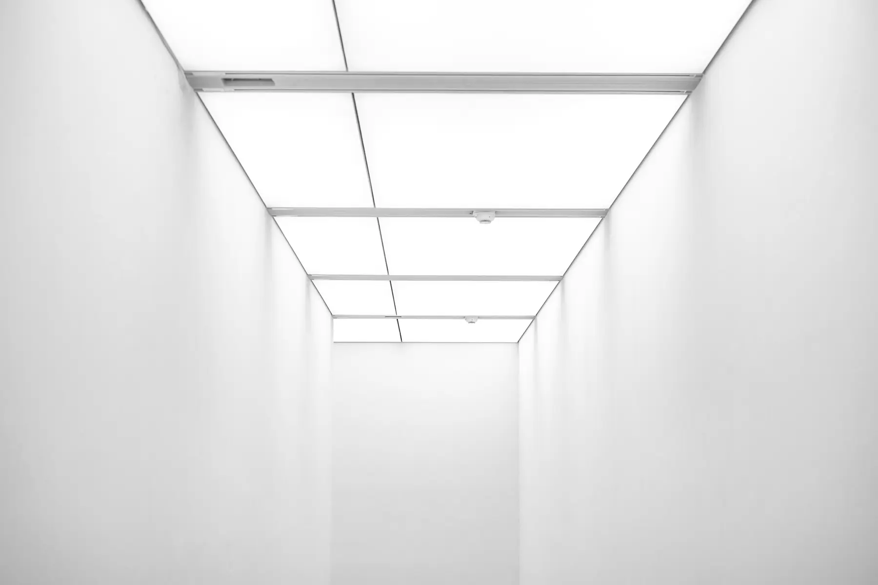 Empty white corridor with luminous ceiling panels receding to a blank wall