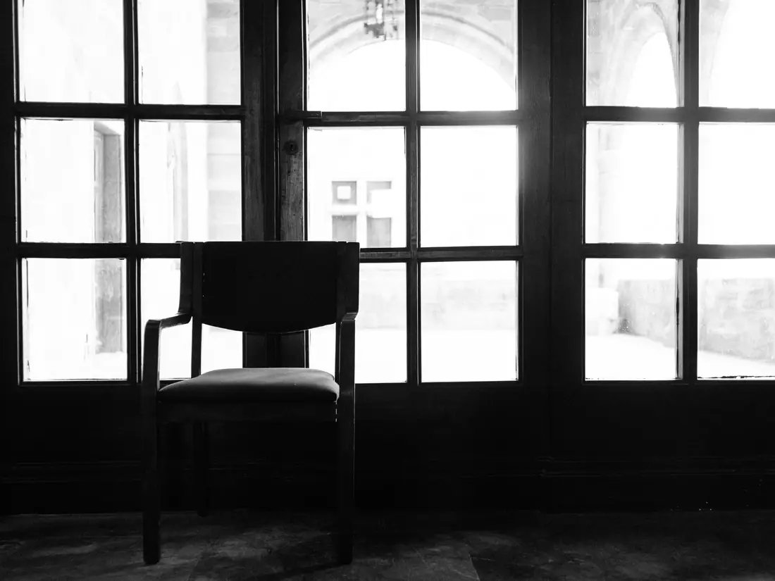 Empty wooden chair silhouetted against tall paned windows full of light