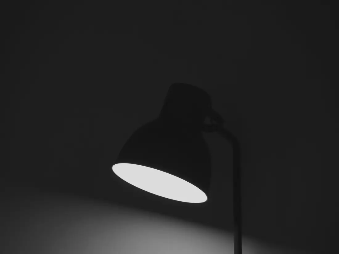 A single black desk lamp lit in a dark room, light pooling below
