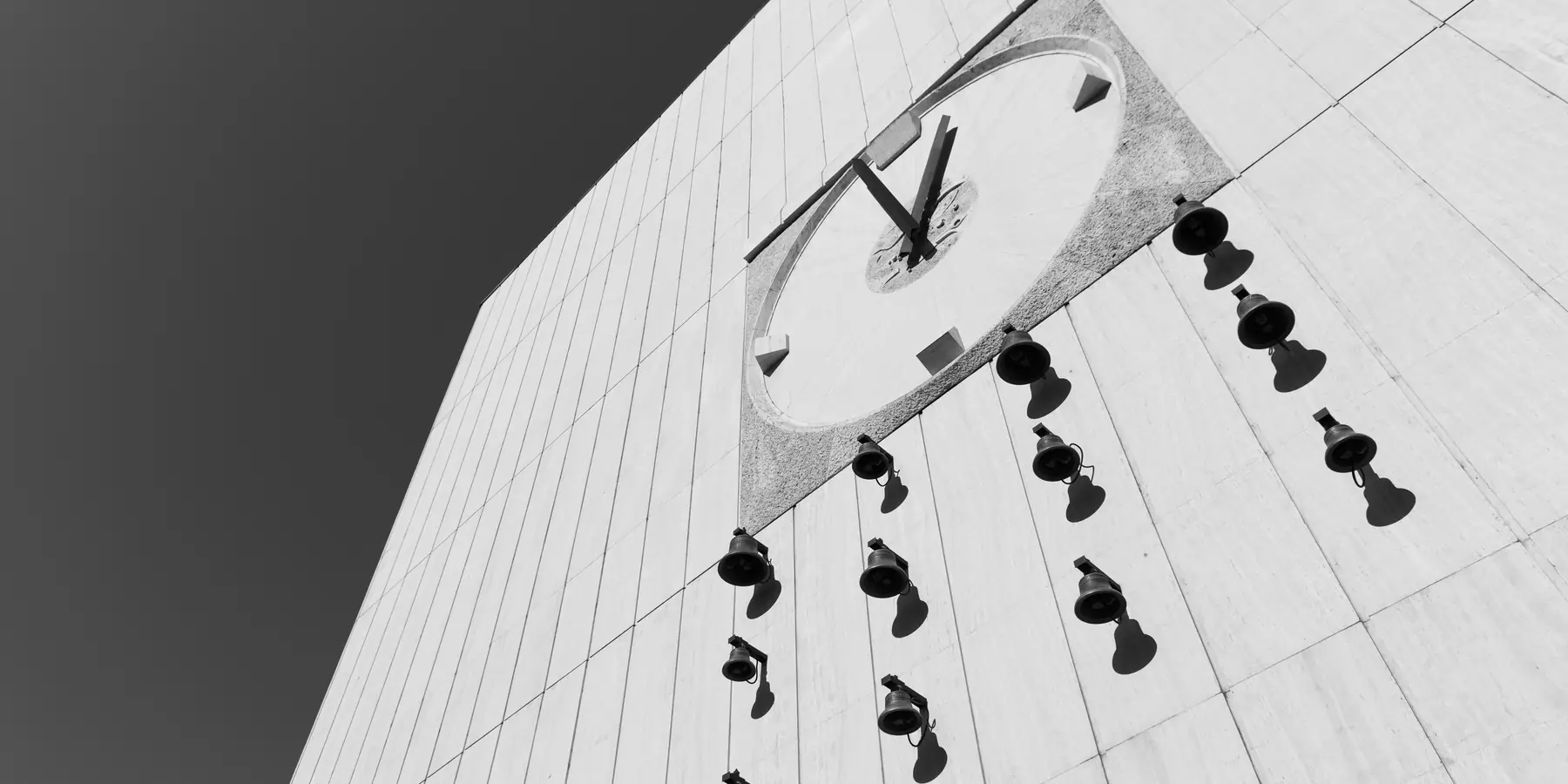 Numeral-free clock on a pale concrete wall, bells and shadows hanging beneath